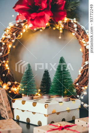 Decorative small Christmas trees on the gift box with elegant flower wreath on gray wall background. Festive home decor. Merry Christmas and New Year. Vertical card with copy space for writing text. Decorative small Christmas trees on the gift box with elegant flower wreath on gray wall background. Festive home decor. Merry Christmas and New Year. Vertical card with copy space for writing text. 93772249