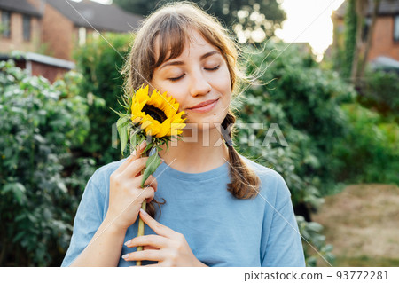Portrait of tender young woman with closed eyes in blue t shirt holding fresh yellow sunflower and enjoying the moment in sunset light on the backyard. Summertime. Selective focus. Copy space. 93772281
