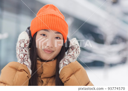 Close-up portrait of a young beautiful Asian woman looking at the camera and smiling while listening to music with big headphones, dressed warmly, on a snowy winter day 93774570