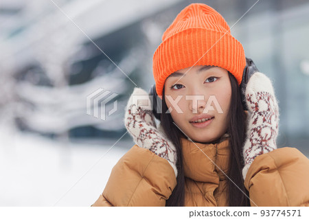 Close-up portrait of a young beautiful Asian woman looking at the camera and smiling while listening to music with big headphones, dressed warmly, on a snowy winter day Close-up portrait of a young beautiful Asian woman looking at the camera and smiling while listening to music with big headphones, dressed warmly, on a snowy winter day 93774571