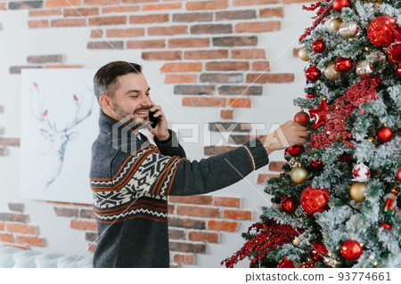 The man talks on the phone and decorates the Christmas tree at home, invites friends to visit, smiles and has fun on Christmas 93774661
