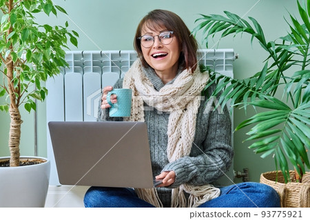 Woman in warm clothes sitting near heating radiator, using laptop 93775921