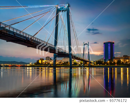 Suspension bridge bridge across the river at night. Night illumination of buildings, reflections. 93776172