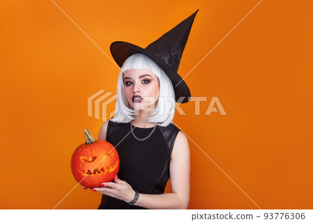 Beautiful sexy woman in witch hat and costume holding carved pumpkin on orange background. Beautiful sexy woman in witch hat and costume holding carved pumpkin on orange background. 93776306