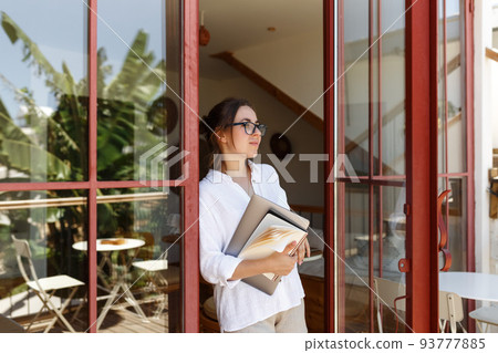 Portrait of businesswoman or student with folder and laptop in hand standing near the glass door background and looking forward. 93777885