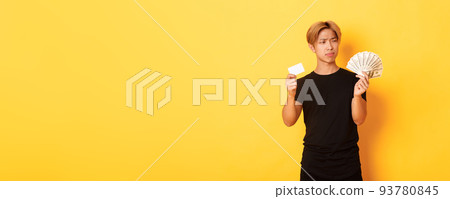 Indecisive and confused young asian guy looking puzzled at money while holding cash and credit card, yellow background 93780845