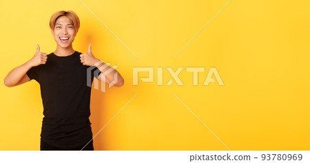 Portrait of satisfied handsome asian male student showing thumbs-up in approval, standing yellow background 93780969