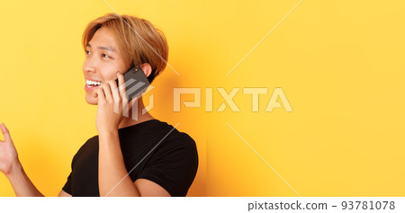 Close-up of attractive korean guy smiling and talking on mobile phone happy, turning left, standing yellow background 93781078