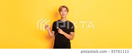 Smiling asian male student with blond hair, pointing fingers right, showing logo, standing yellow background Smiling asian male student with blond hair, pointing fingers right, showing logo, standing yellow background 93781113