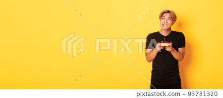 Portrait of handsome dreamy asian guy looking upper left corner and thinking, making wish while celebrating birthday and holding b-day cake, yellow background 93781320