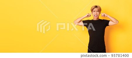 Portrait of annoyed and bothered asian guy grimacing, shut ears disturbed by loud noise, standing yellow background 93781400