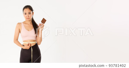 Sport, wellbeing and active lifestyle concept. Disappointed gloomy asian girl measuring waist with tape measure and sulking as cannot eat chocolate bar while losing weight on diet 93781402