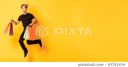 Full-length of happy attractive asian man jumping from happiness and carry shopping bags, standing yellow background Full-length of happy attractive asian man jumping from happiness and carry shopping bags, standing yellow background 93781434