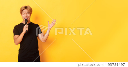Handsome asian male singer, korean guy singing song at karaoke in microphone with passion, standing over yellow background Handsome asian male singer, korean guy singing song at karaoke in microphone with passion, standing over yellow background 93781497
