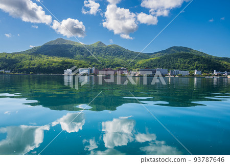 The surface of Lake Toya reflects the blue sky, white clouds, Mt. Usu, and the hot spring town like a mirror 93787646