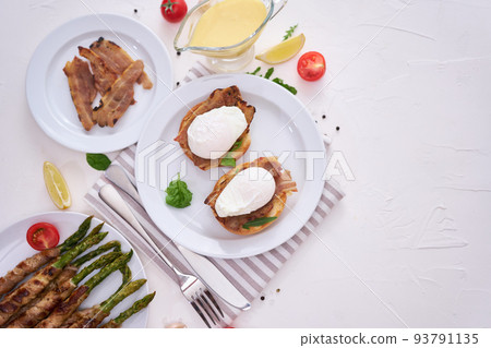 Tasty eggs Benedict, hollandaise sauce and aspargus covered with bacon 93791135