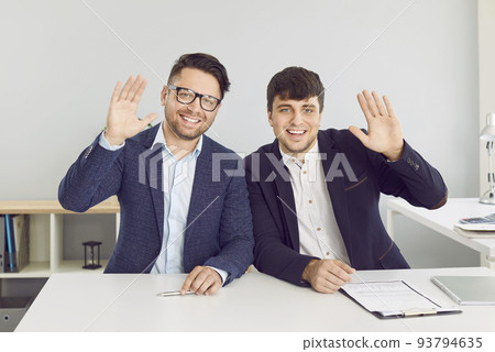 Two happy business people sitting in office, smiling, and waving hello at online conference Two happy business people sitting in office, smiling, and waving hello at online conference 93794635