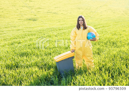 A woman in a yellow protective suit stands in the middle of a green field holding a globe, and with her other hand carries a garbage can. The concept of saving the planet from environmental pollution A woman in a yellow protective suit stands in the middle of a green field holding a globe, and with her other hand carries a garbage can. The concept of saving the planet from environmental pollution 93796309