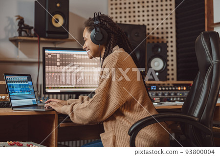 Charming woman blogger, anchorwoman, typing text on laptop, records online podcast using headphones and microphone in professional music studio 93800004