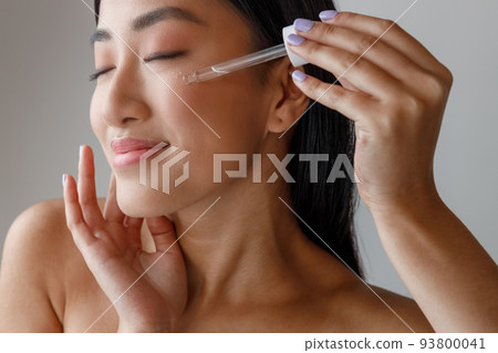 Smiling Asian woman applying vitamin serum on her face 93800041