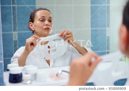Reflection in bathroom mirror of a beautiful dark-haired multi-ethnic woman, in white bathrobe applying a smoothing moisturizing textile face mask. Skin care. Home spa. Anti-aging beauty procedures 93800909
