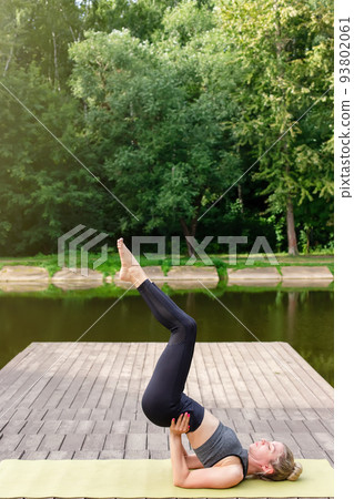 A slim woman on a wooden platform by a pond in a park in summer, does yoga A slim woman on a wooden platform by a pond in a park in summer, does yoga 93802061