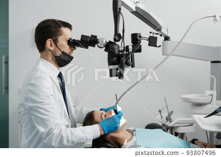 Male dentist using dental microscope in dental office with female patient 93807496