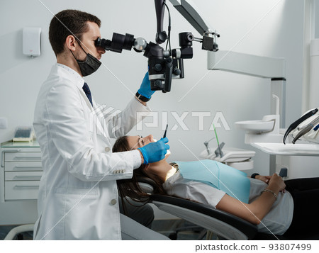 Male dentist using dental microscope in dental office with female patient 93807499