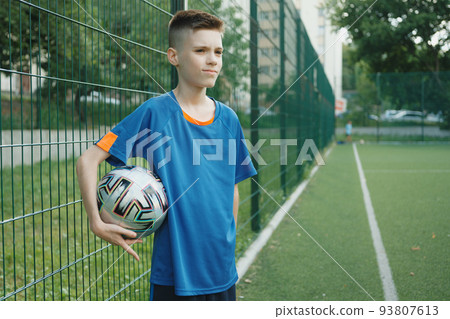 Photo of confident teenage boy in blue sportswear holding soccer ball on the play field 93807613