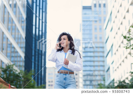 Young business woman walking across the road with tablet among the skyscrapers and speaks on the phone. 93808743