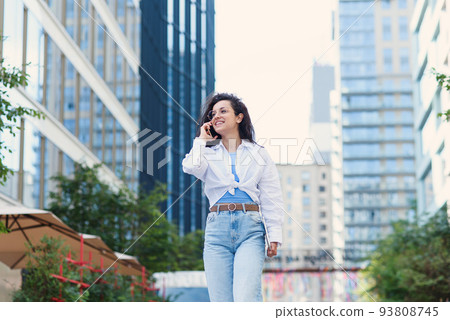Young business woman walking across the road with tablet among the skyscrapers and speaks on the phone. 93808745