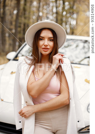 Portrait of woman in autumn forest, standing in front of her white car. Brunette woman posing for a photo. Woman wearing white hat, pink t-shirt and trousers. 93814908