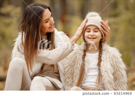 Young woman with little girl sitting on a fallen tree trunk in autumn forest. Brunette woman play with her daughter. Girl wearing beige sweater and mother wearing white clothes. 93814969