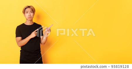 Portrait of disappointed and sad handsome blond asian guy, sulking upset, pointing fingers upper right corner, standing yellow background 93819128