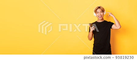 Happy satisfied asian guy likes music or podcast, pointing finger at headphones, holding smartphone, standing yellow background 93819230