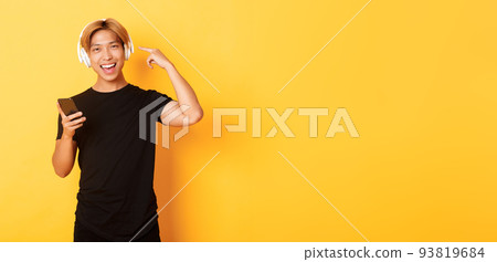 Happy satisfied asian guy likes music or podcast, pointing finger at headphones, holding smartphone, standing yellow background 93819684