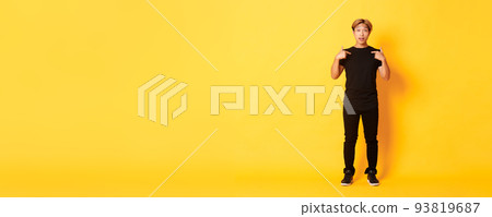 Full-length of surprised attractive korean blond guy in black outfit pointing at himself impressed, yellow background 93819687