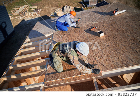 Carpenters hammering nail into OSB panel on the roof top of future cottage. Men workers building wooden frame house. Carpentry and construction concept. 93822737