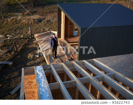 Male architect building wooden frame house in the Scandinavian style barnhouse. Man standing on terrace at construction site, inspecting quality of work in the evening. 93822743