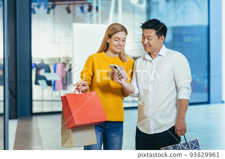 Diverse couple Asian man and blonde woman in clothes supermarket, looking at mobile phone screen, choosing shopping, holding colorful shopping bags and gifts. 93829961