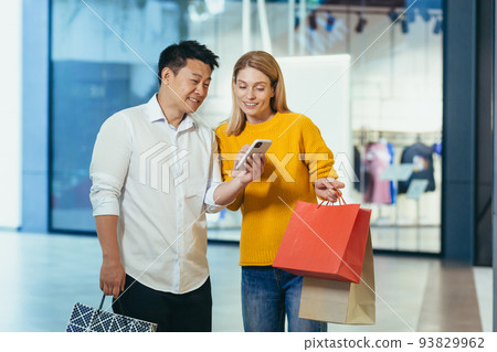 Diverse couple Asian man and blonde woman in clothes supermarket, looking at mobile phone screen, choosing shopping, holding colorful shopping bags and gifts. 93829962