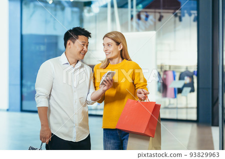 Diverse couple Asian man and blonde woman in clothes supermarket, looking at mobile phone screen, choosing shopping, holding colorful shopping bags and gifts. 93829963