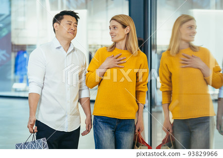 multiracial couple asian man and caucasian woman walking together in mall after shopping with colored bags in hands. walking past shop windows with clothes cheerful family hugging smile. multiracial couple asian man and caucasian woman walking together in mall after shopping with colored bags in hands. walking past shop windows with clothes cheerful family hugging smile. 93829966