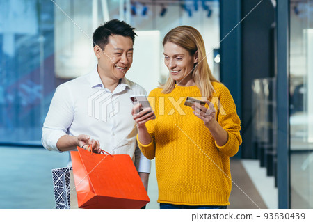 Diverse couple of shoppers Asian man and woman shopping for clothes inside store smiling and happy holding bank credit card and colorful shopping Diverse couple of shoppers Asian man and woman shopping for clothes inside store smiling and happy holding bank credit card and colorful shopping 93830439