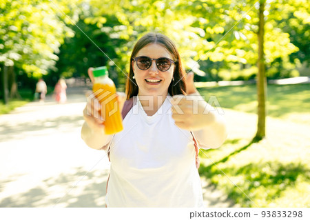 Young stylish happy woman with juice giving thumb up as sign of success and standing over nature background. Redhead girl walking in the park and holding a bottle of fresh cold smoothie in a sunny day 93833298
