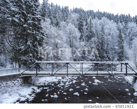 snow-covered pedestrian bridge over Elbe or Labe river and spruce tree forest at ski resort Spindleruv mlyn, Krkonose mountains, Czech republic. Winter day. snow-covered pedestrian bridge over Elbe or Labe river and spruce tree forest at ski resort Spindleruv mlyn, Krkonose mountains, Czech republic. Winter day. 93851921