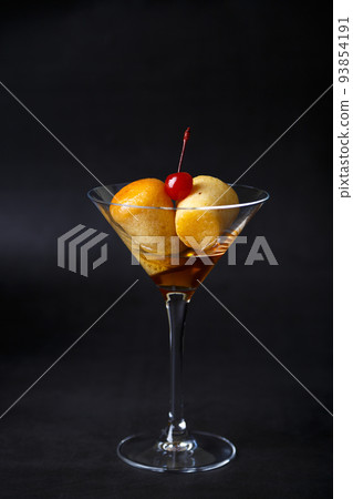 Neapolitan Rum baba (or baba au rum) in a martini glass with a cocktail cherry on a black background. Small yeast cakes soaked in rum syrup. Traditional Italian pastry. Close-up, selective focus. Neapolitan Rum baba (or baba au rum) in a martini glass with a cocktail cherry on a black background. Small yeast cakes soaked in rum syrup. Traditional Italian pastry. Close-up, selective focus. 93854191