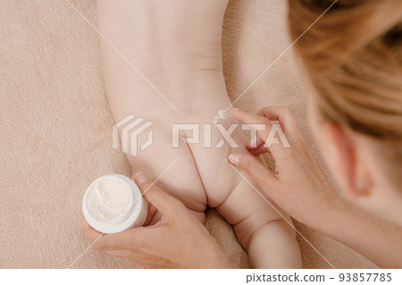 Baby is lying on a white sheets. Baby bum and feet. Mom applying some cream. Close-up of mother hands applying cream under the diaper on little baby pop. Baby is lying on a white sheets. Baby bum and feet. Mom applying some cream. Close-up of mother hands applying cream under the diaper on little baby pop. 93857785