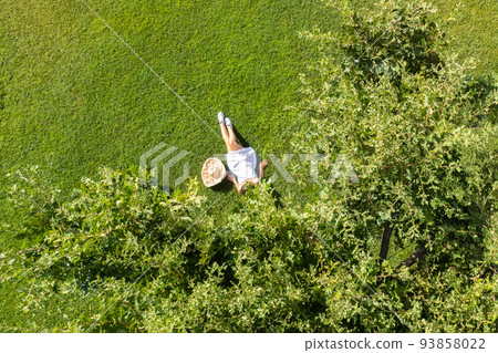 Woman in a white dress, hat with laptop, lying on the green grass under tree. Top view, drone, aerial view 93858022