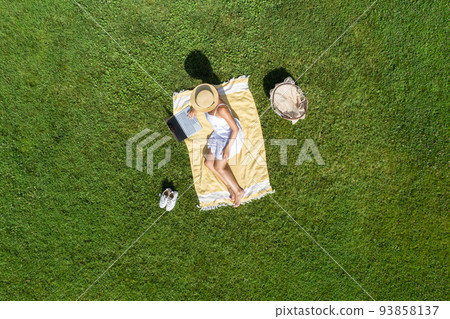 Woman in a white dress and hat, sitting on the plaid on the green grass having a picnic with food basket. Top view, drone, aerial view 93858137